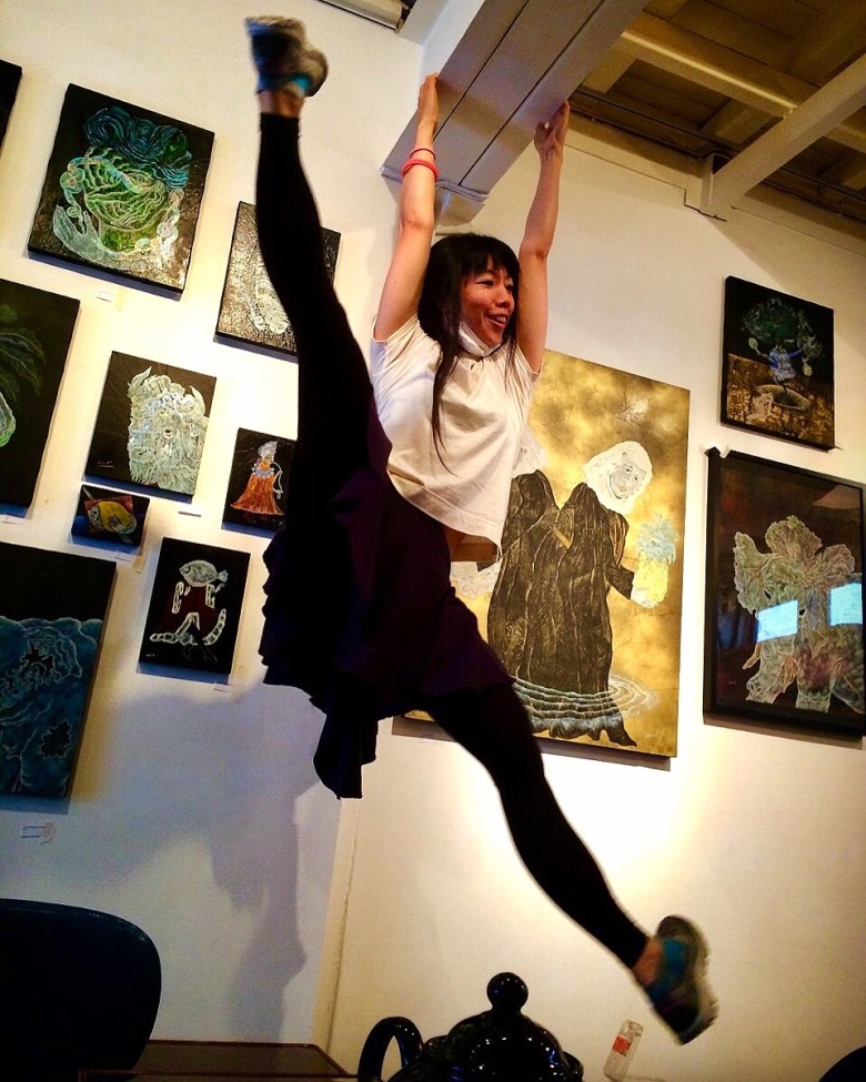 Mako Kubeck dancing while hanging by her arms from a rafter in front of artwork by HAMADARAKA at the Tetoka gallery